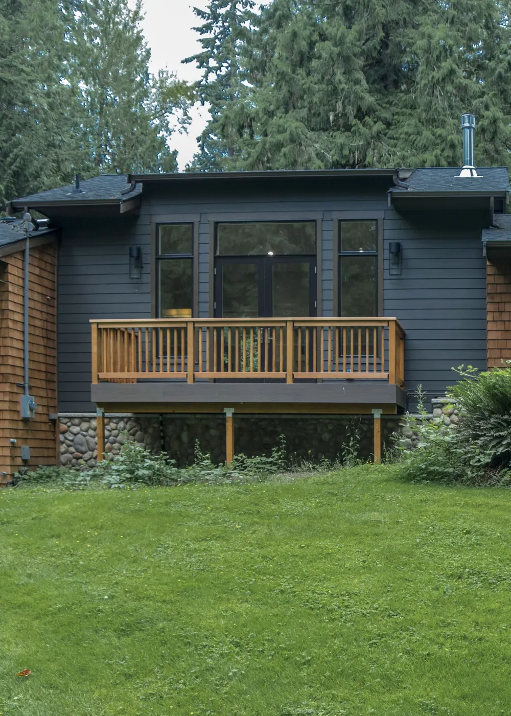 Dark two-story house with a deck and lawn, surrounded by tall trees